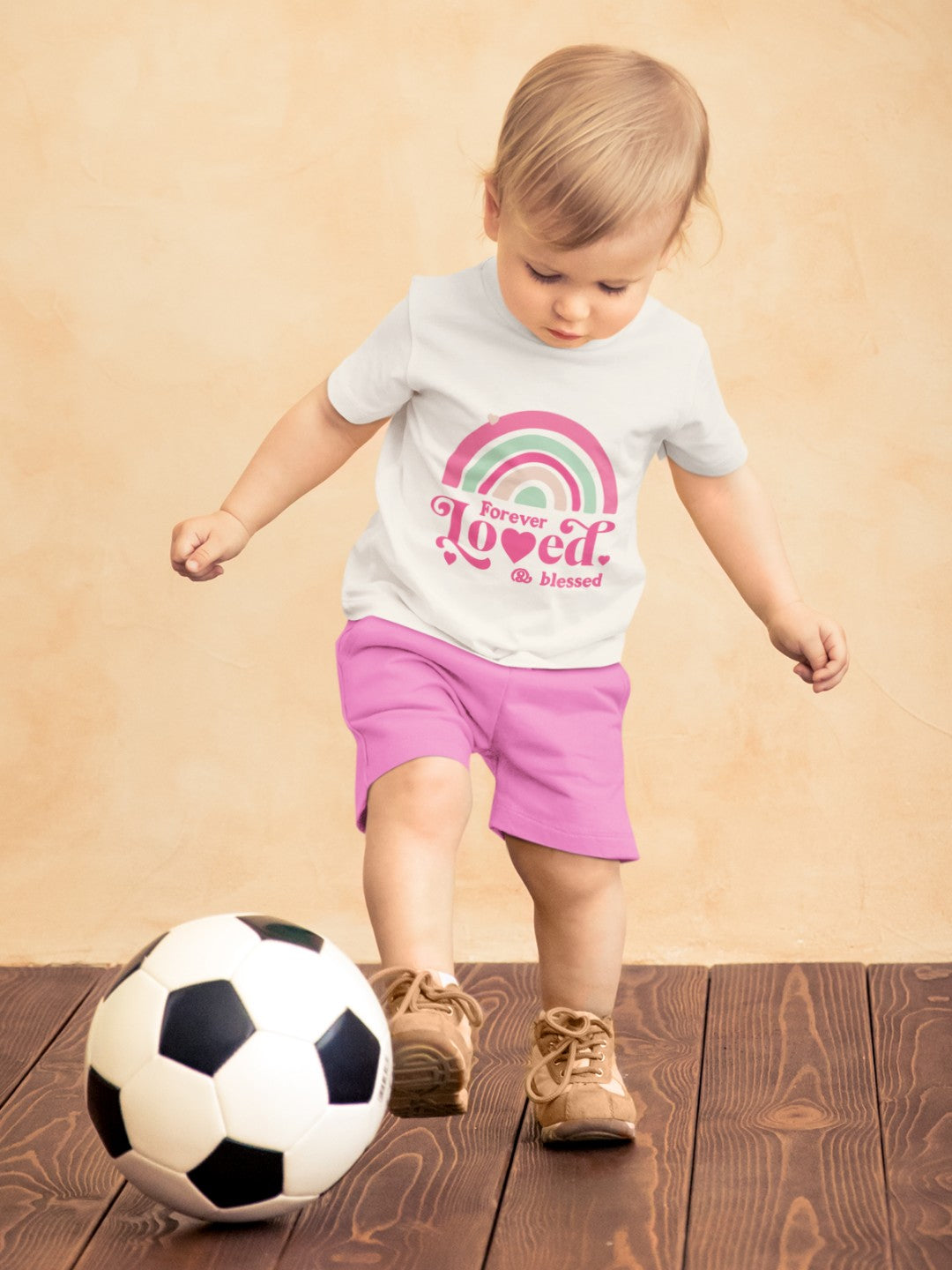 Toddler wearing cute rainbow toddler t-shirt with pink shorts playing with a soccer ball